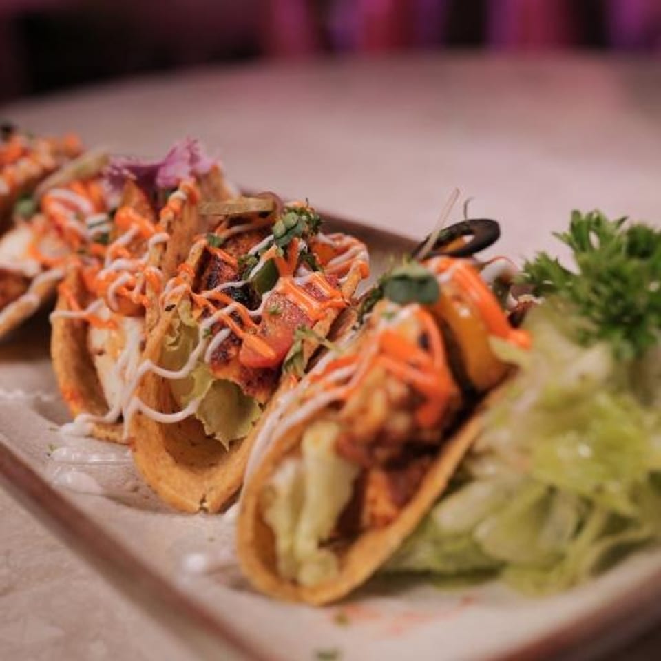 Three crispy tacos topped with grilled seafood, shredded vegetables, creamy sauce, herbs, and lettuce garnish on a rectangular plate.